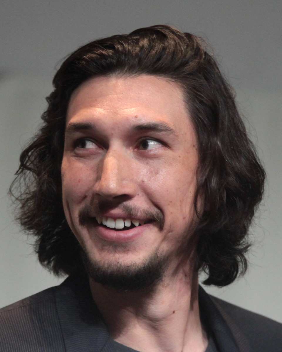 Adam Driver laughing, with shoulder-length dark wavy hair and a goatee, in a dark jacket