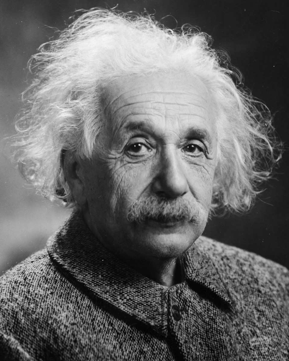 Albert Einstein in black and white, with wild white hair and a bushy mustache, in a tweed jacket, looking straight at the camera