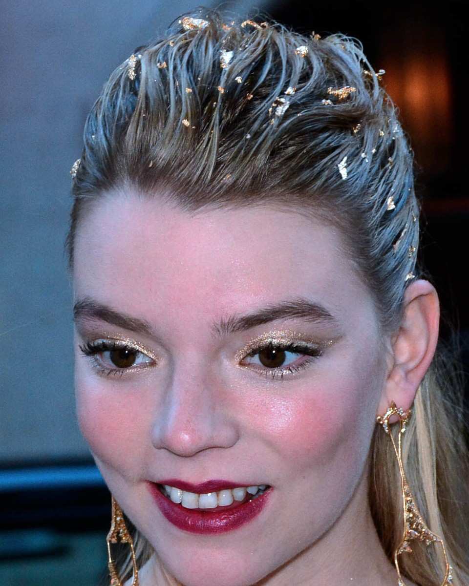 Anya Taylor-Joy smiling with slicked-back blonde hair flecked with gold and gold drop earrings