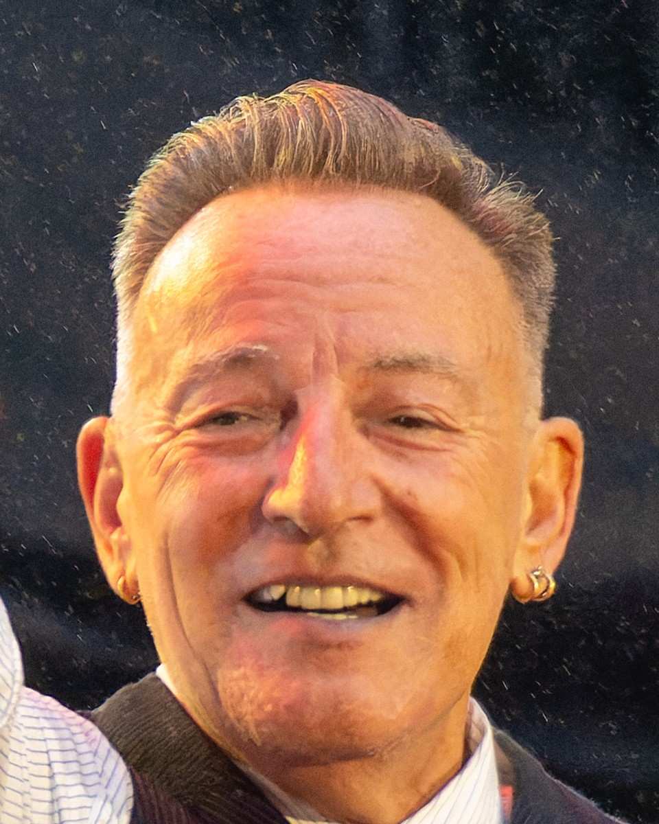 Bruce Springsteen smiling on stage in a striped shirt, vest and tie, a gold hoop earring in each ear