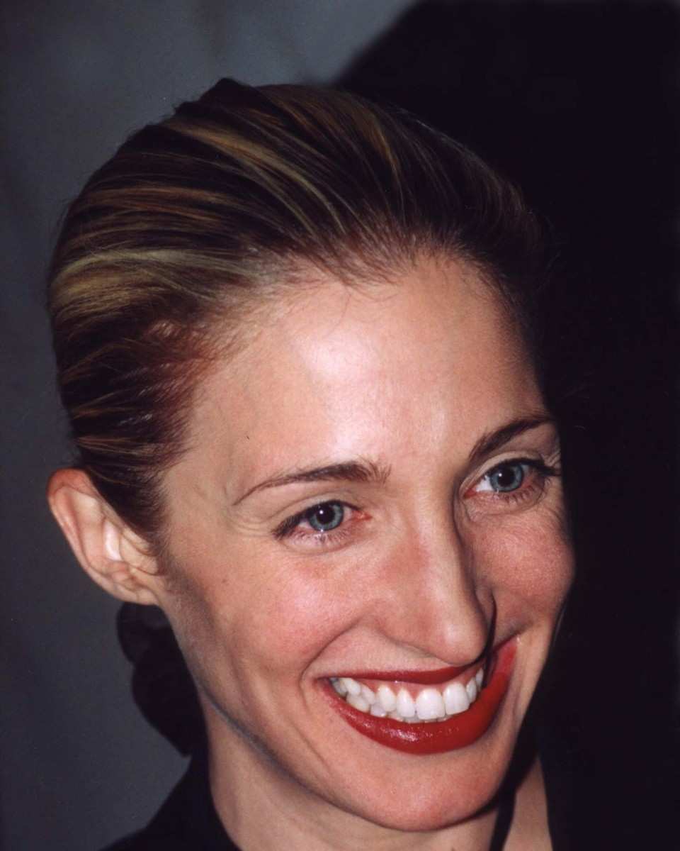 Carolyn Bessette-Kennedy smiling broadly with her hair slicked back and red lipstick, in a dark top with crossed straps