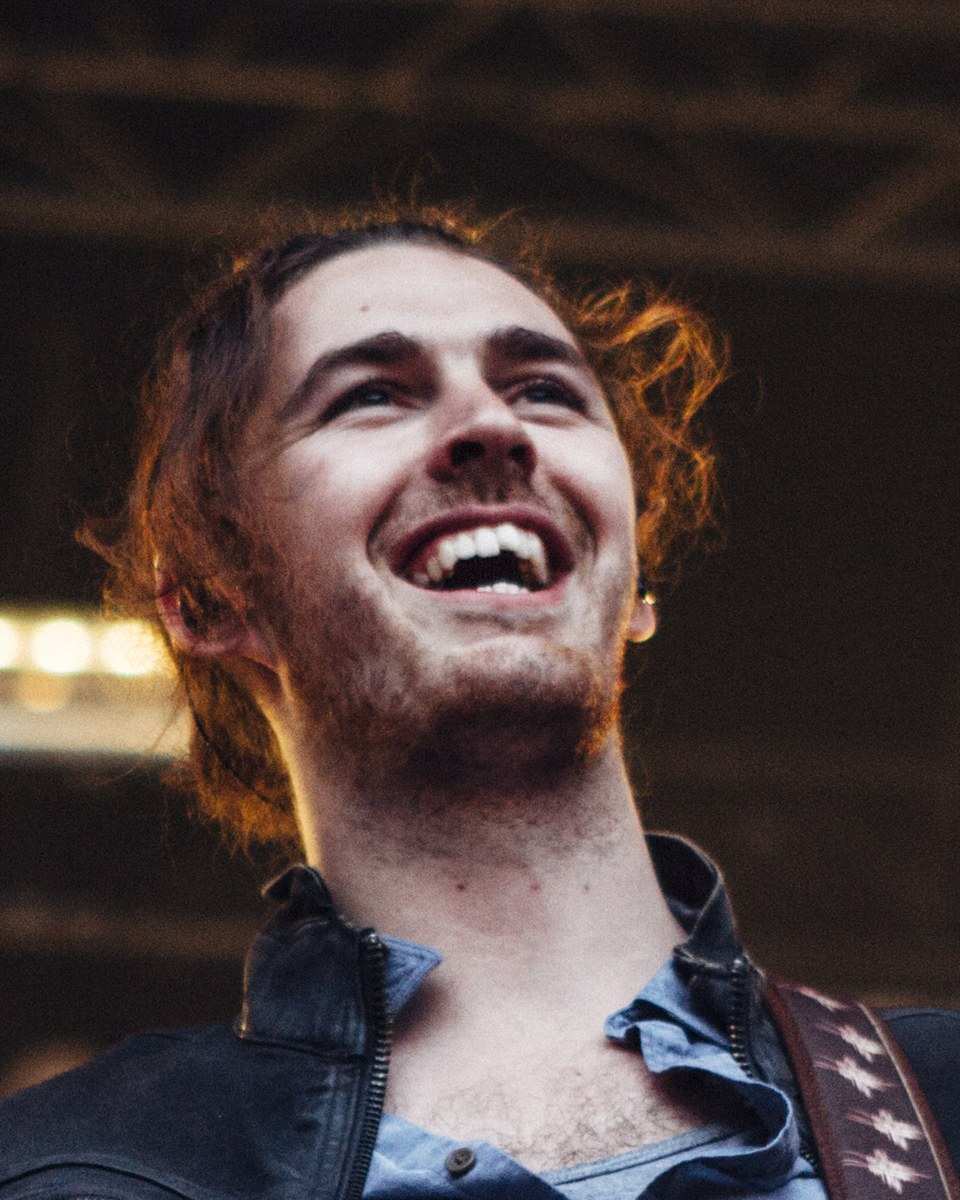 Hozier laughing on an outdoor stage in a black jacket over a denim shirt