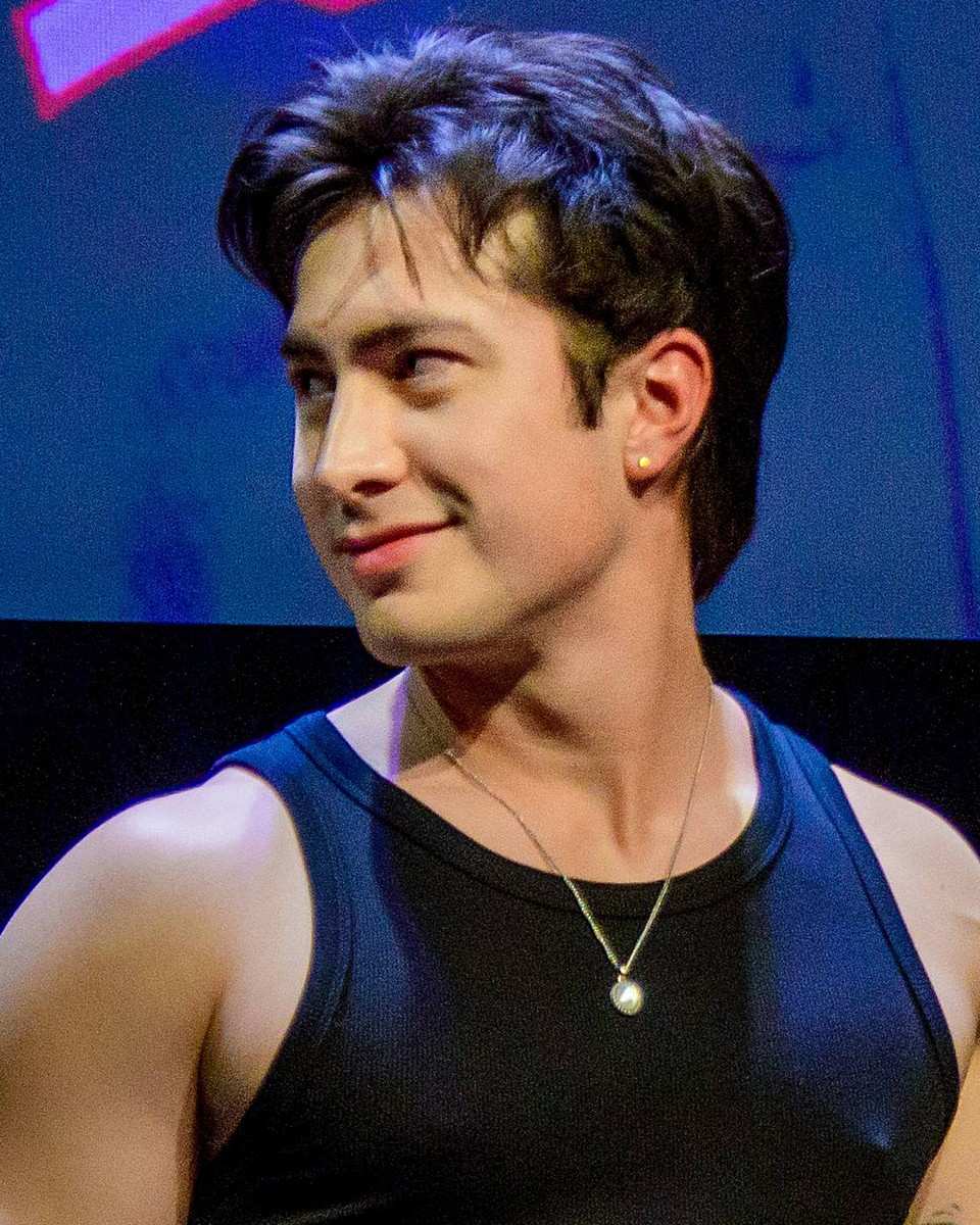 Hudson Williams with dark swept-up wavy hair, a stud earring and a pendant necklace, glancing to the side with a faint smirk, in a dark tank top