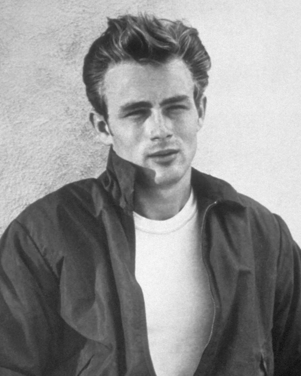 James Dean in a dark zip-up jacket over a white T-shirt, leaning against a wall, in black and white