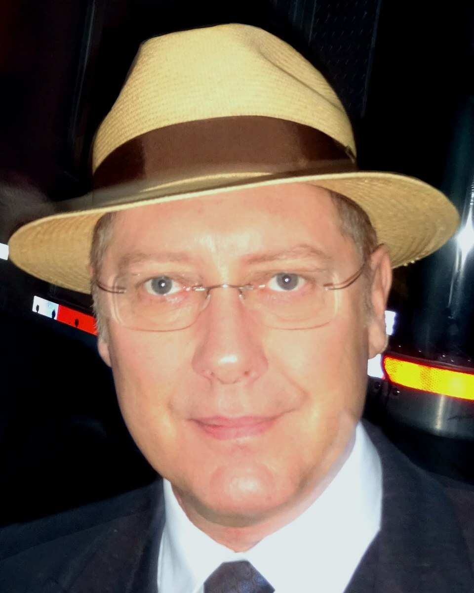 James Spader in a straw fedora, glasses and a dark suit
