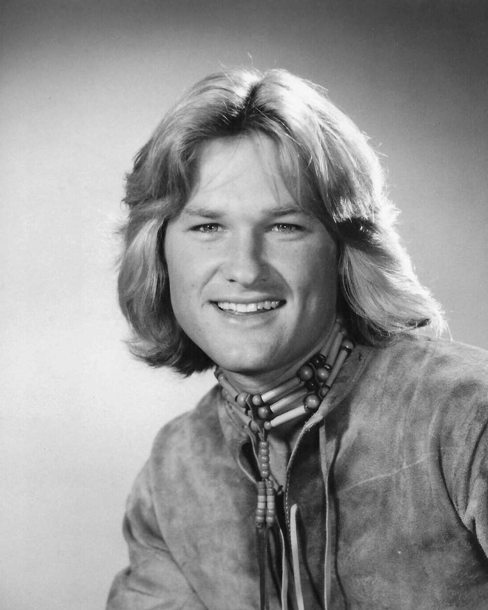 Kurt Russell smiling with feathered shoulder-length hair, a suede jacket and a beaded necklace, in black and white