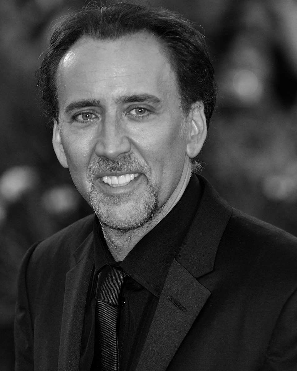 Nicolas Cage in a black suit and tie with a goatee, in black and white