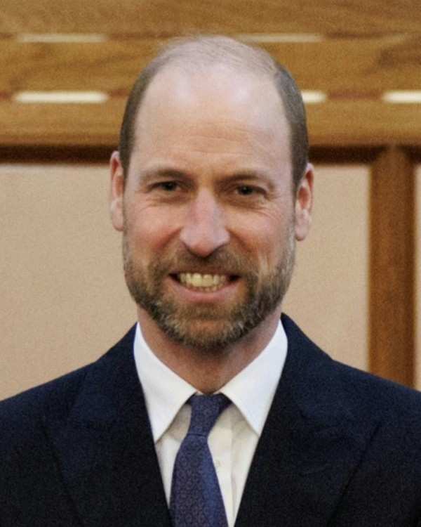 Prince William smiling in a dark navy suit, white shirt and patterned blue tie, in front of wood panelling
