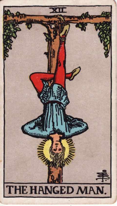 The Hanged Man card of the Rider–Waite–Smith deck: a man in a blue tunic and red tights hangs upside down by one foot from a T-shaped wooden beam with leaves, his other leg bent behind the knee, his hands behind his back and a halo around his head