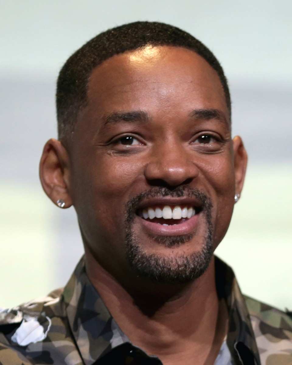 Will Smith in a close portrait, smiling broadly with a goatee, a stud earring in each ear and a camouflage-pattern collared shirt