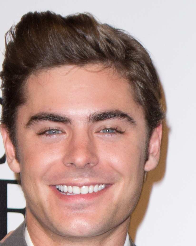 Zac Efron smiling in a grey suit and striped tie at a premiere